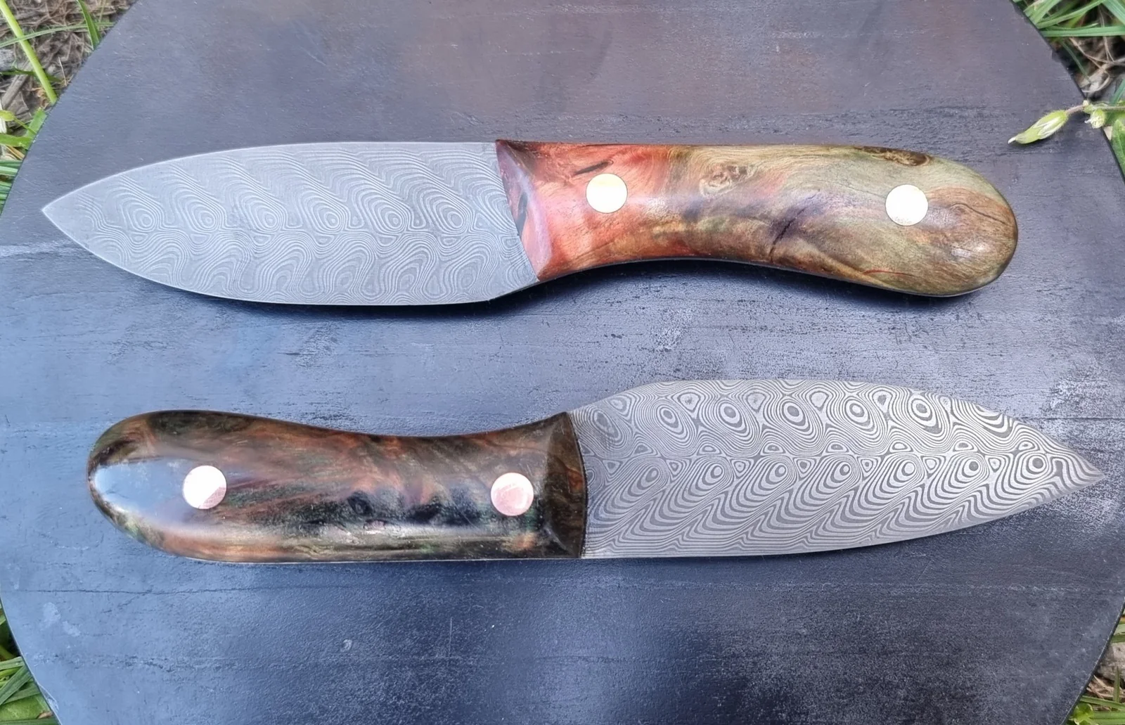Two knives with patterned blades and wooden handles on a dark surface.